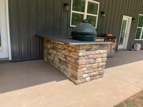Stacked stone outdoor kitchen island with a grill and ceramic cooker on a covered porch